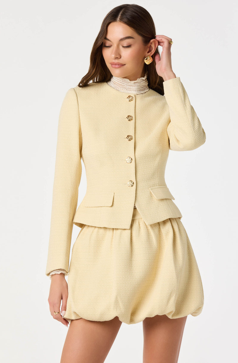 A woman is wearing a cream-colored, textured set consisting of a fitted, long-sleeved blazer with gold buttons and two flap pockets, paired with matching puffed shorts. She is also wearing a high-neck, pleated, striped blouse underneath the blazer, and accessorizing with gold earrings and a ring.