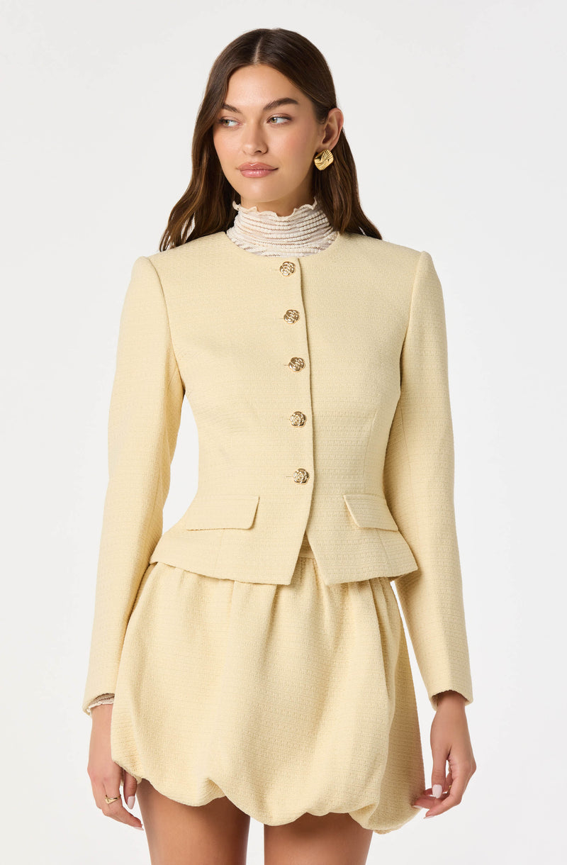 A woman is wearing a cream-colored, textured blazer with long sleeves and a high neckline, paired with a matching skirt that has a puffy, bubble-style shape. The blazer features gold buttons down the front and small flap pockets at the waist. She accessorizes with large, gold, textured earrings.