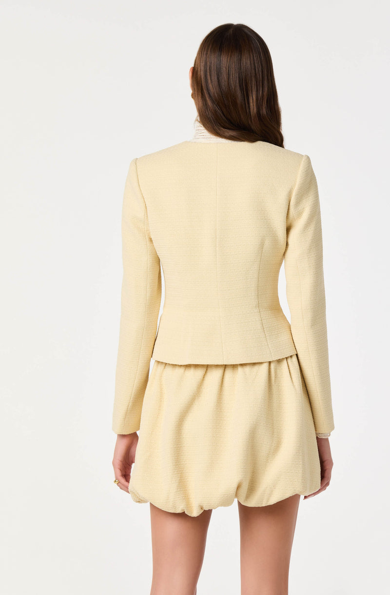 A woman is wearing a light-colored, tailored blazer with structured shoulders and long sleeves. The matching shorts are loose-fitting with a gathered, elastic waistband and a bubble hem.