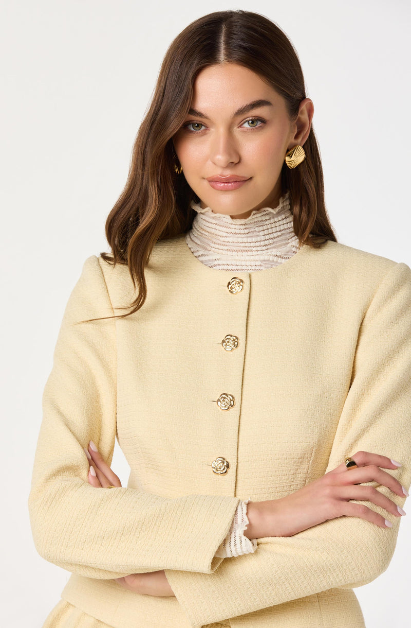 A woman is wearing a light yellow textured jacket with a rounded neckline and gold decorative buttons down the front. She has accessorized with large, gold, sculptural earrings and a white ribbed turtleneck underneath the jacket.