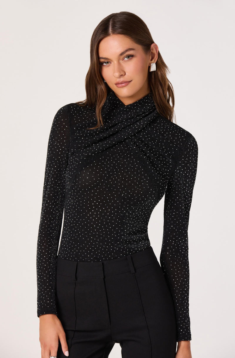 A woman is wearing a long-sleeved, fitted black top with a high, draped neckline. The top is decorated with small, evenly spaced white polka dots throughout. She has paired it with high-waisted black trousers.