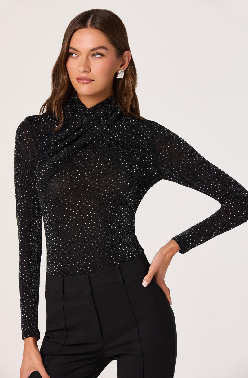 A woman is wearing a fitted, long-sleeved black top adorned with small white dots throughout. The top has a high neckline that drapes slightly, creating a gathered effect around the neck and shoulders. She is also wearing high-waisted black pants with a tailored fit. The woman has minimal makeup and shoulder-length brown hair styled in loose waves. She accessorizes with a pair of large, square, silver earrings.