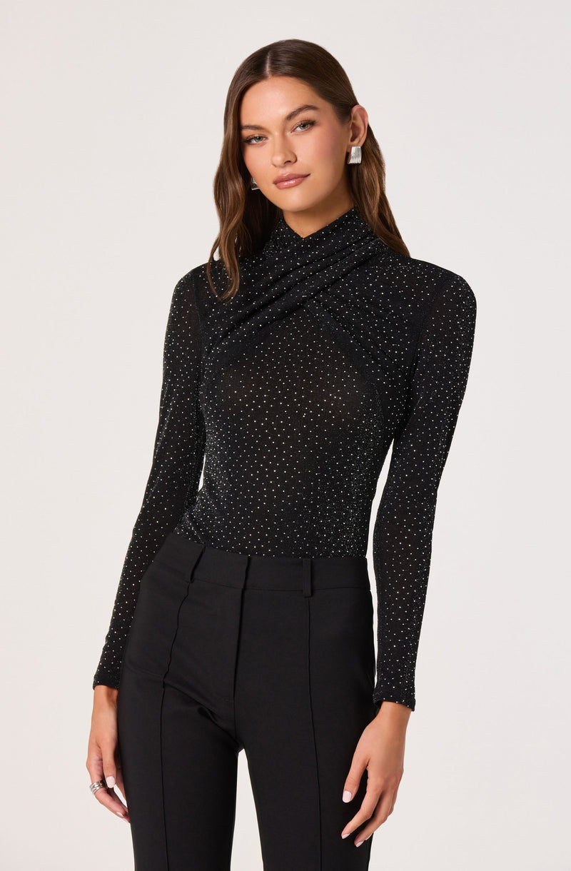 A woman is wearing a black long-sleeve top with a high neckline that features a pattern of small white dots, creating a sparkling effect. She pairs it with high-waisted black trousers that have a tailored fit and visible seam lines running down the front.
