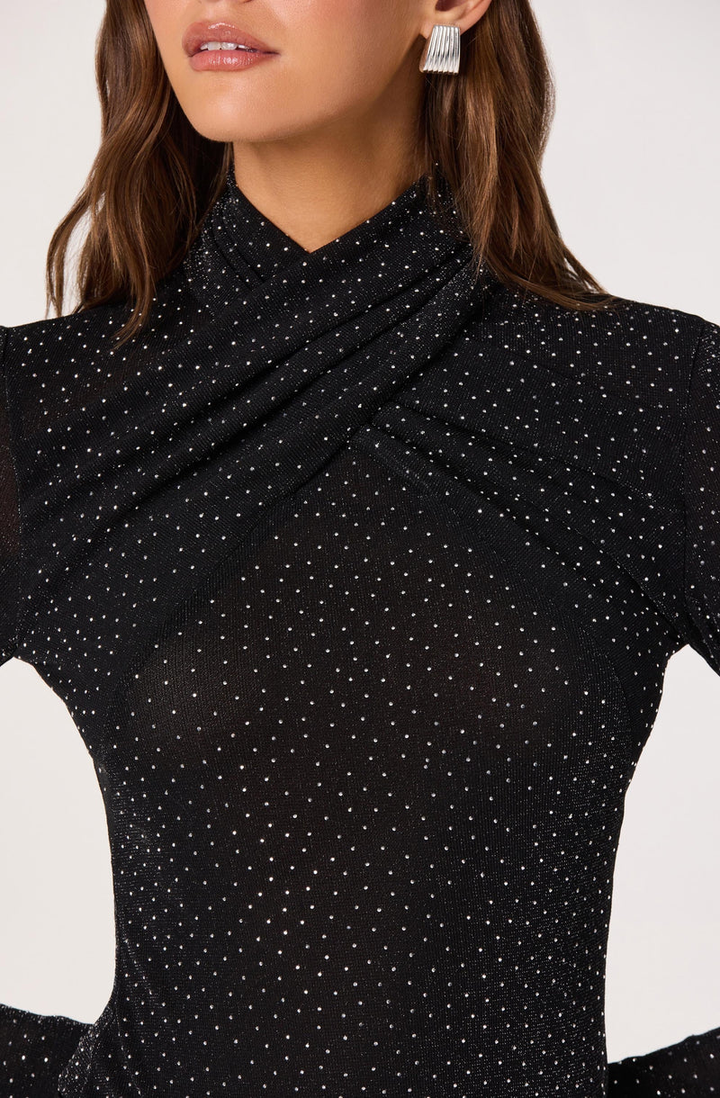 A woman is wearing a black, long-sleeved top embellished with small, shiny silver dots that create a sparkling pattern. The top features a high neckline with a draped, gathered design around the shoulders and chest area. She is accessorized with large, rectangular, silver earrings.