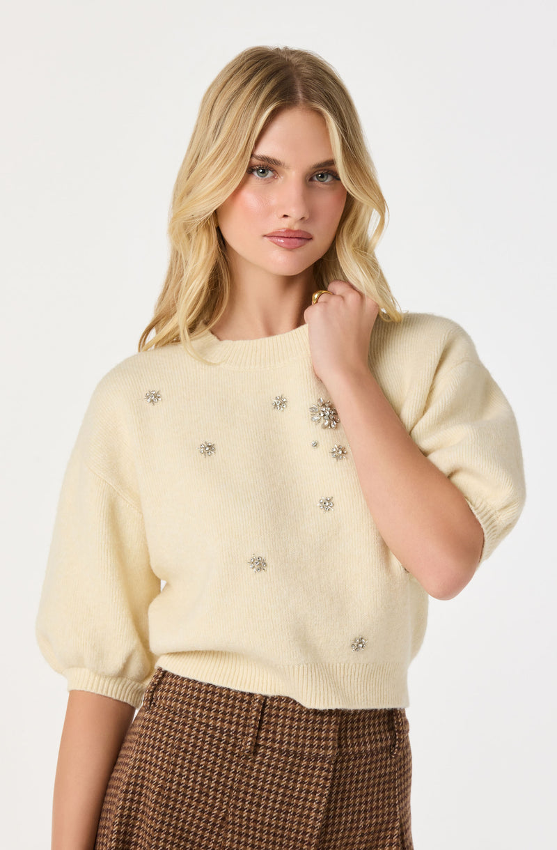 A woman is wearing a cream-colored knit sweater with a round neckline and short, puffed sleeves. The sweater is decorated with small, silver, floral-shaped embellishments scattered across the front. She is also wearing high-waisted, brown checkered pants.