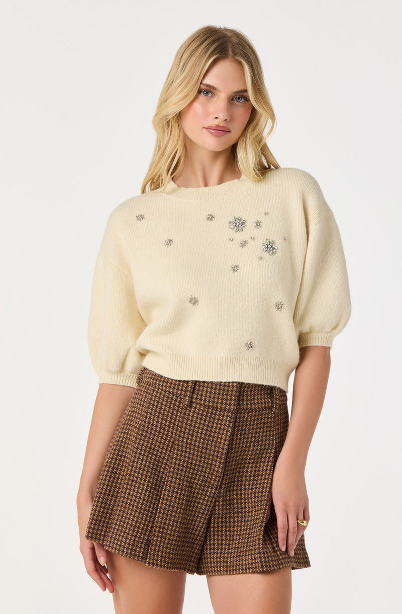 A woman is wearing a cream-colored sweater featuring decorative silver floral embellishments scattered across the front. The sweater has a relaxed fit with slightly puffed sleeves that are gathered at the cuffs. She pairs the sweater with high-waisted, brown houndstooth patterned shorts.