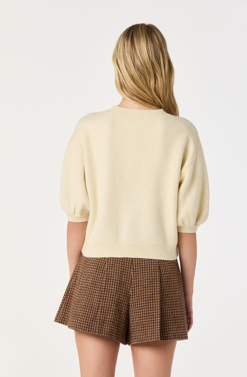A person is wearing a cream-colored, short-sleeved knit sweater with puffed sleeves that are gathered at the cuffs. They are also dressed in high-waisted, brown plaid shorts with a pleated design. The background is plain white.