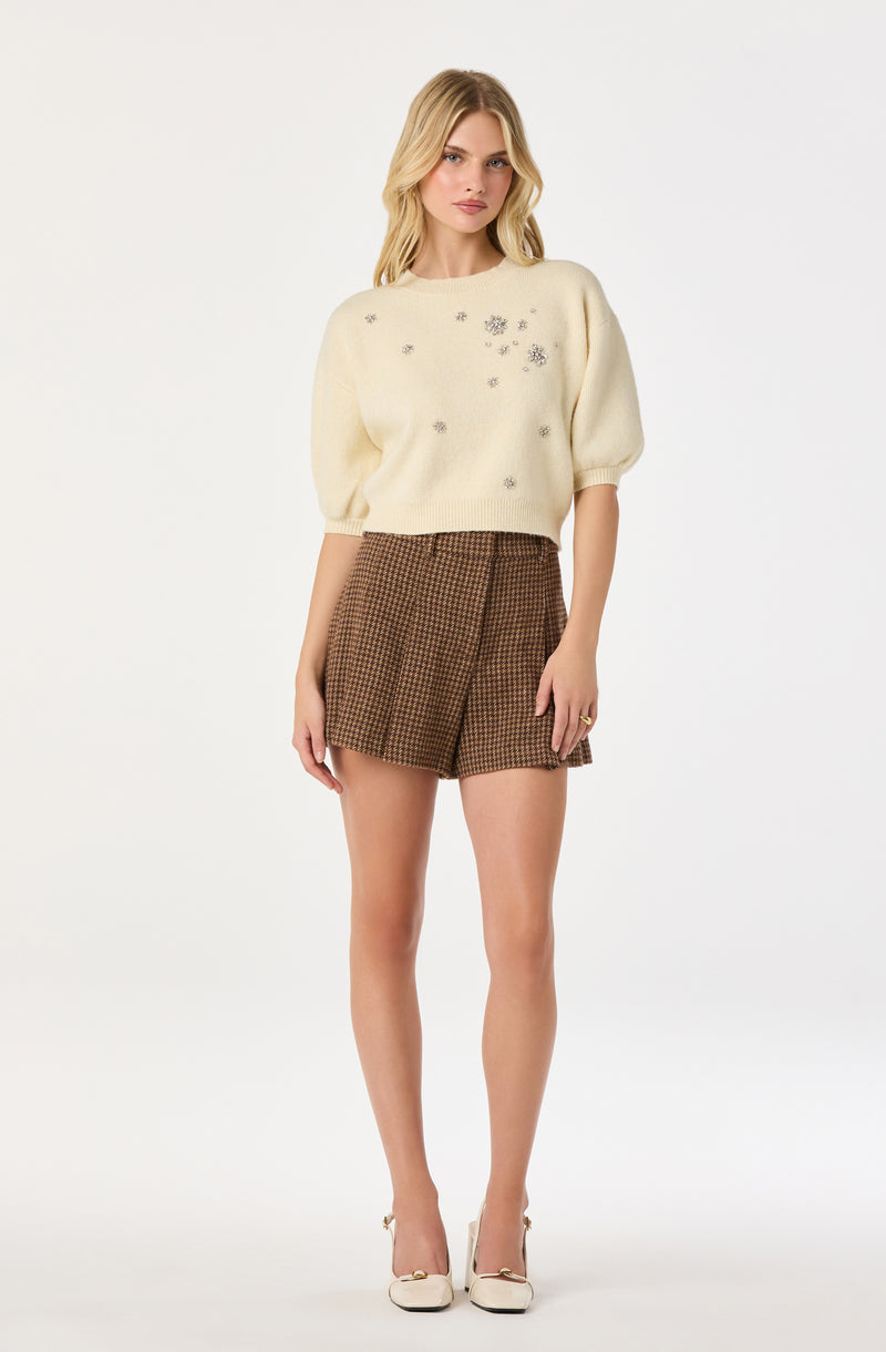 A woman is wearing a cream-colored, loose-fitting sweater with short puffed sleeves and metallic floral embellishments on the front. She has paired it with high-waisted, brown plaid shorts and white high-heeled shoes.