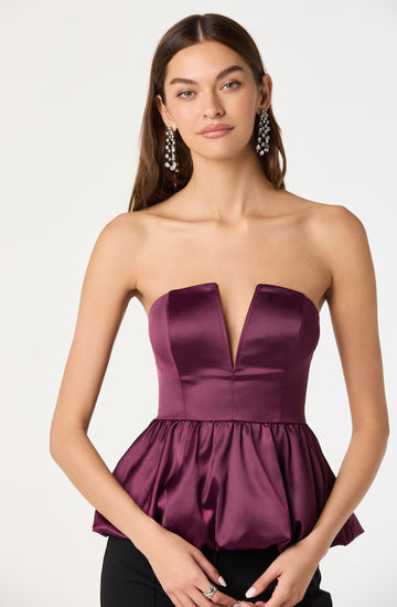 A woman is wearing a strapless, deep purple satin top with a plunging V-neckline and a fitted bodice. The bottom of the top features a gathered, bubble-style design. She is accessorized with large, dangling, crystal-embellished earrings.