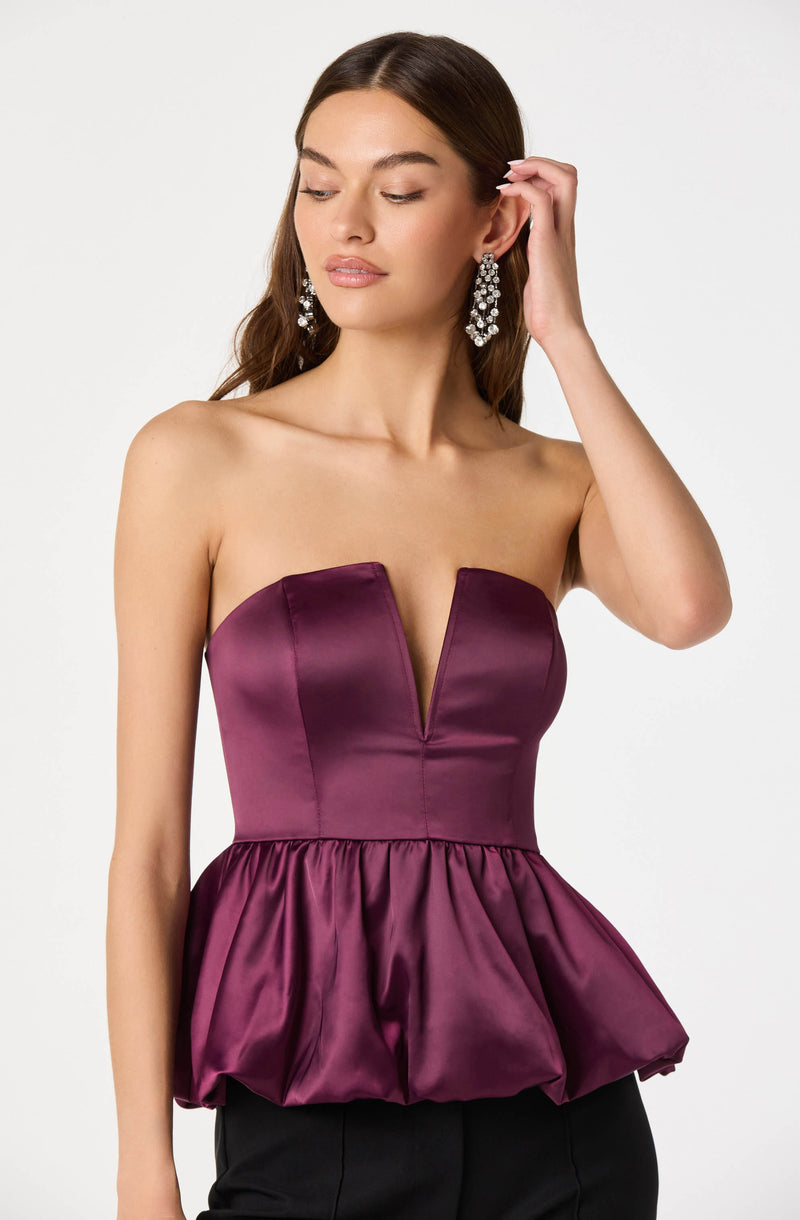 A woman is wearing a strapless, deep purple satin top with a V-shaped neckline, fitted at the waist with a gathered, bubble-style peplum. She has long, wavy brown hair and is accessorized with large, dangling, crystal statement earrings.