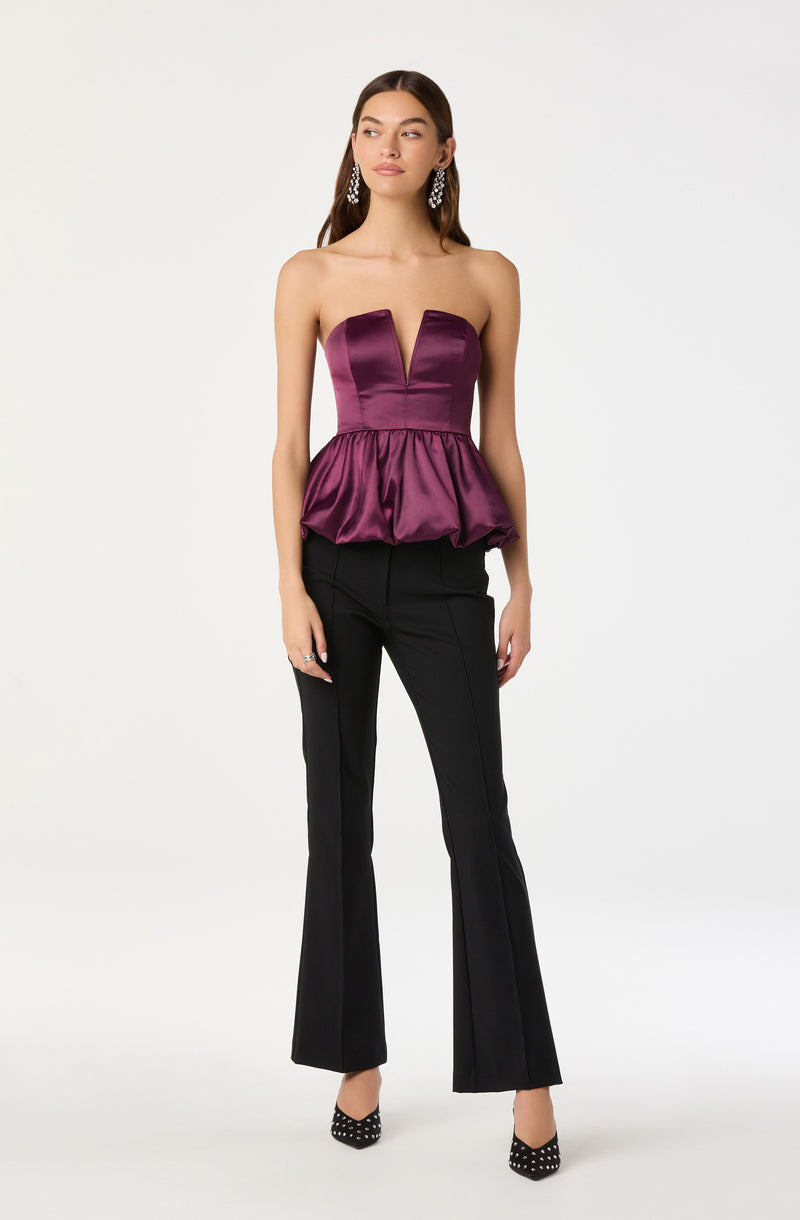 A woman is wearing a strapless, deep purple satin top with a plunging neckline and a gathered peplum at the waist. She pairs it with black, high-waisted, flared pants. She is accessorized with long, dangling earrings and black high heels with white polka dots.