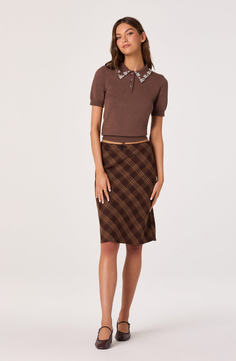 A woman is wearing a brown short-sleeved sweater with a collared neckline decorated with white floral embroidery along the edges. She pairs it with a brown plaid knee-length skirt with diagonal lines in shades of brown, yellow, and orange. She completes the outfit with matching brown shoes with ankle straps.