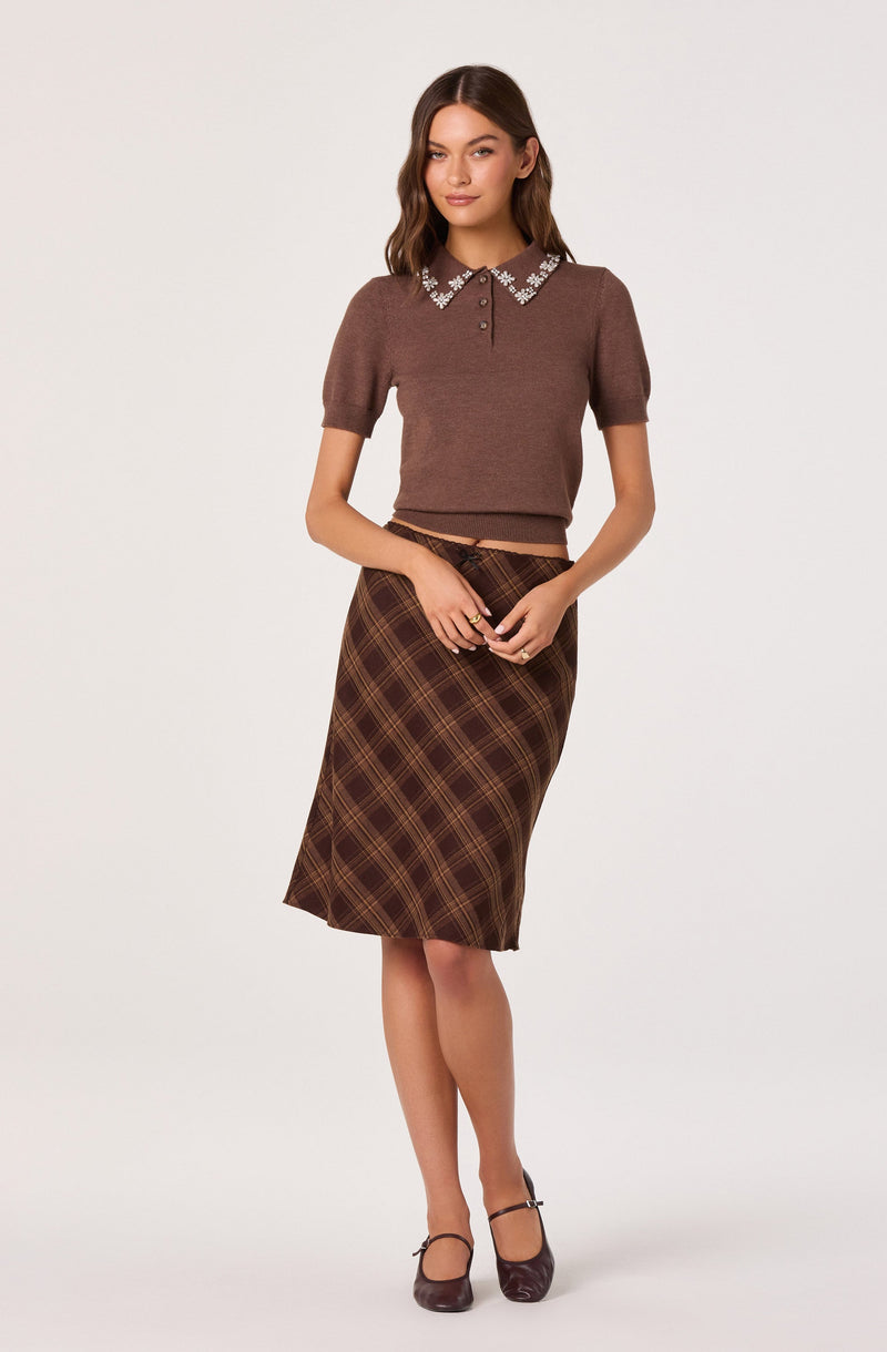 A woman is wearing a brown short-sleeved knit top with a pointed collar decorated with floral embellishments. She is also wearing a high-waisted, knee-length plaid skirt in shades of brown and yellow. She has on dark brown Mary Jane style shoes with a strap across the top.
