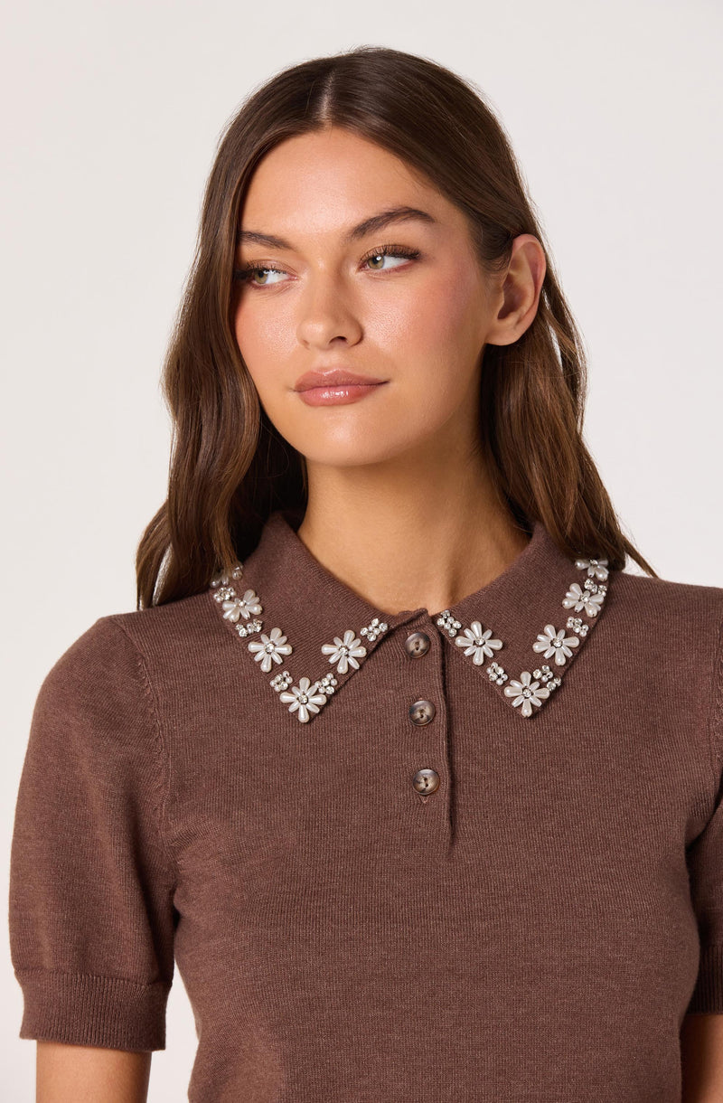 A woman is wearing a brown short-sleeved knit top with a rounded collar decorated with white pearl and rhinestone floral embellishments. The top has a row of three buttons below the collar.