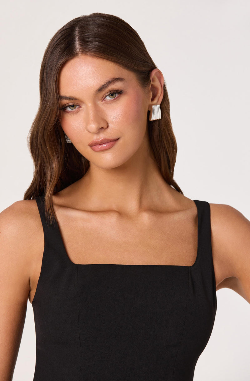 A woman with long, wavy brown hair wearing a sleeveless black dress with wide straps and a square neckline. She is accessorized with large rectangular white earrings.
