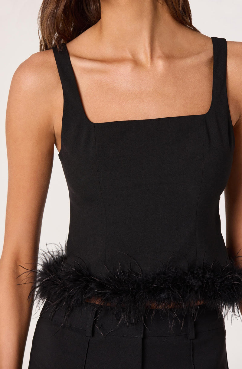 A woman is wearing a black sleeveless top with wide shoulder straps and a square neckline. The top features decorative black feathers around the hemline at the waist.