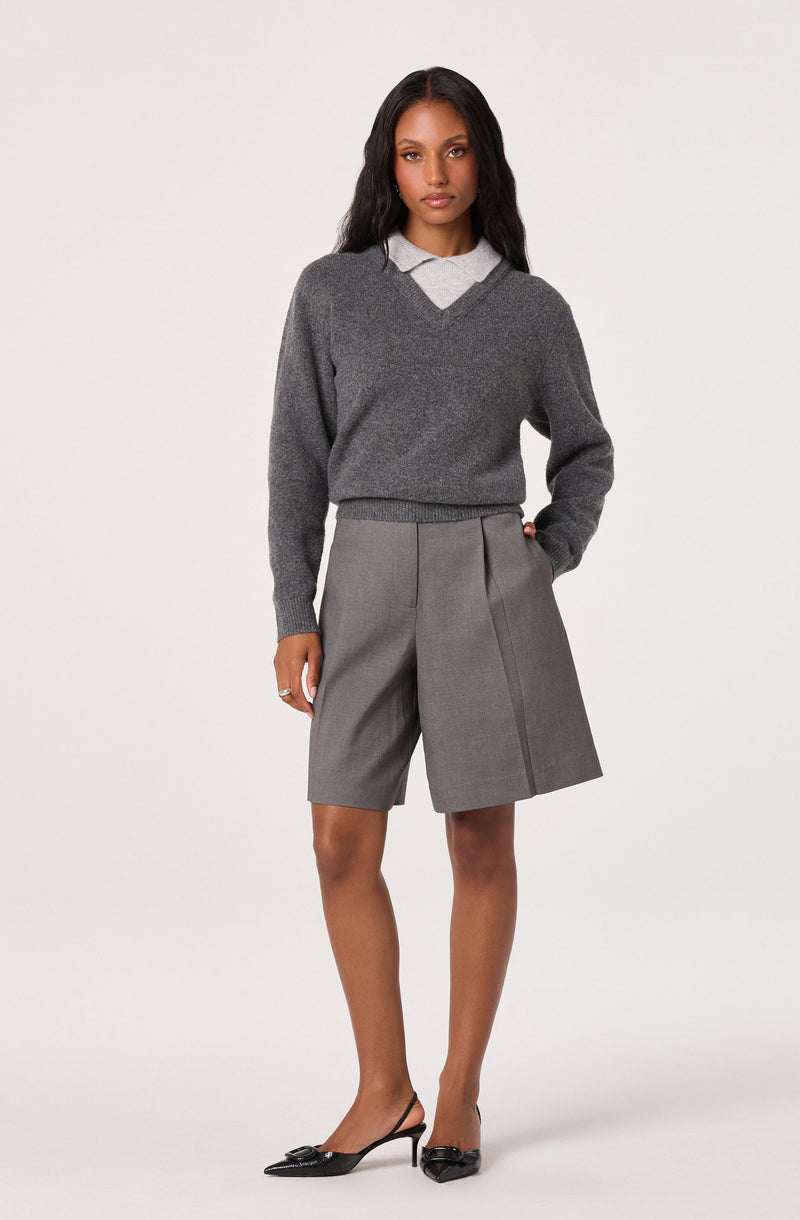 A woman is wearing a dark gray sweater with a V-neckline layered over a light gray collared shirt. She pairs this with high-waisted, knee-length gray shorts that have front pleats and a side seam detail. She completes her outfit with black pointed-toe high heels featuring a bow embellishment on the front.