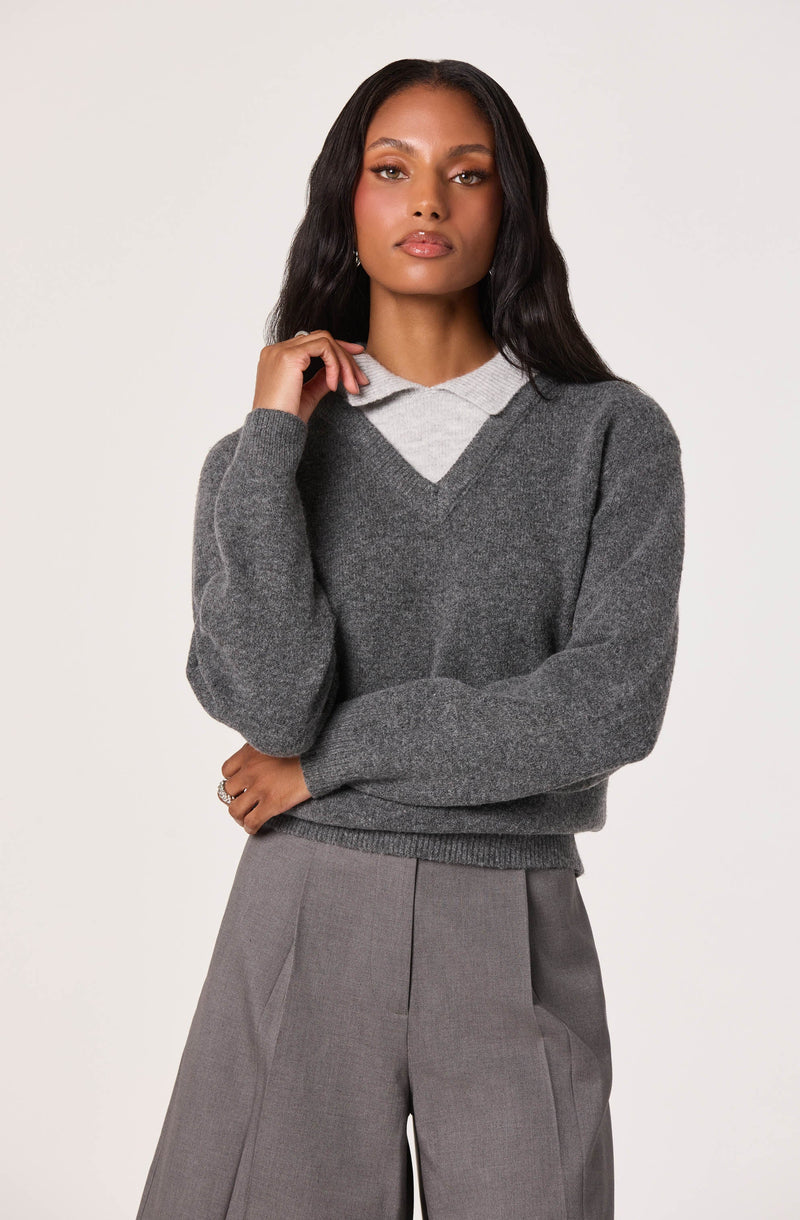 A woman is wearing a dark gray V-neck sweater layered over a light gray collared shirt, paired with high-waisted gray trousers.