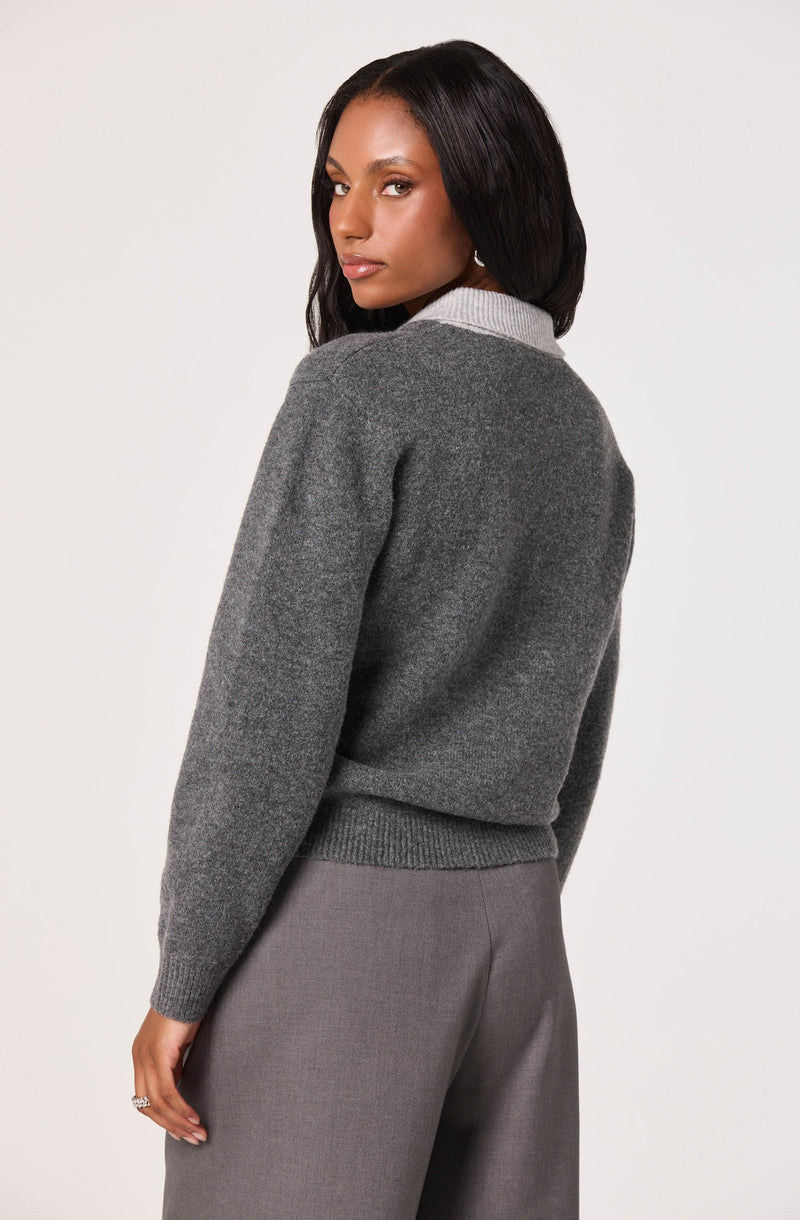 A woman is wearing a gray wool sweater with a light gray collar, paired with gray dress pants. The sweater has a ribbed hem and cuffs, and the overall look is elegant and professional. The woman has long, dark hair and is posed against a plain, light-colored background.