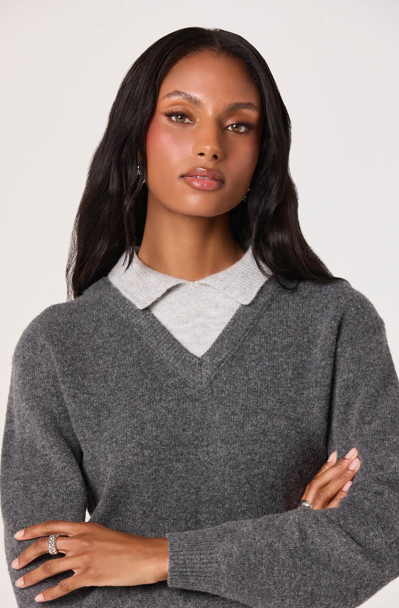 A woman is wearing a layered outfit consisting of a light gray collared shirt underneath a dark gray V-neck sweater.