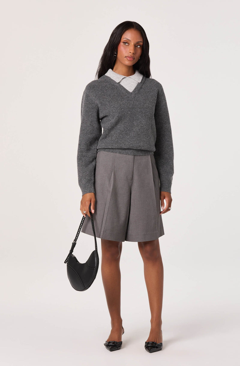 A woman dressed in a light gray collared shirt layered with a dark gray V-neck sweater, paired with knee-length gray pleated shorts. She holds a black handbag with a chain strap in her right hand and wears black pointed-toe heels.