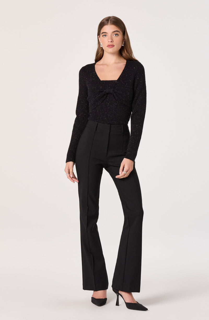 The woman is wearing a black, sparkly long-sleeve top with a square neckline and a decorative bow detail in the front. She is also dressed in high-waisted black trousers with a straight-leg cut, and black high-heeled shoes.