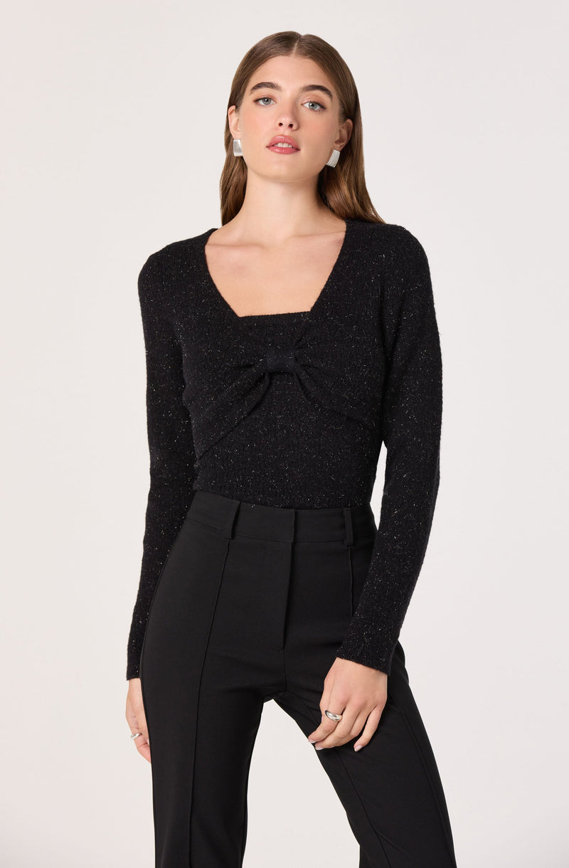 A woman is wearing a black long-sleeve top with a subtle sparkle effect and a large bow detail at the front. She pairs it with high-waisted black trousers that have a tailored fit. She accessorizes with white earrings and a ring on her right hand.
