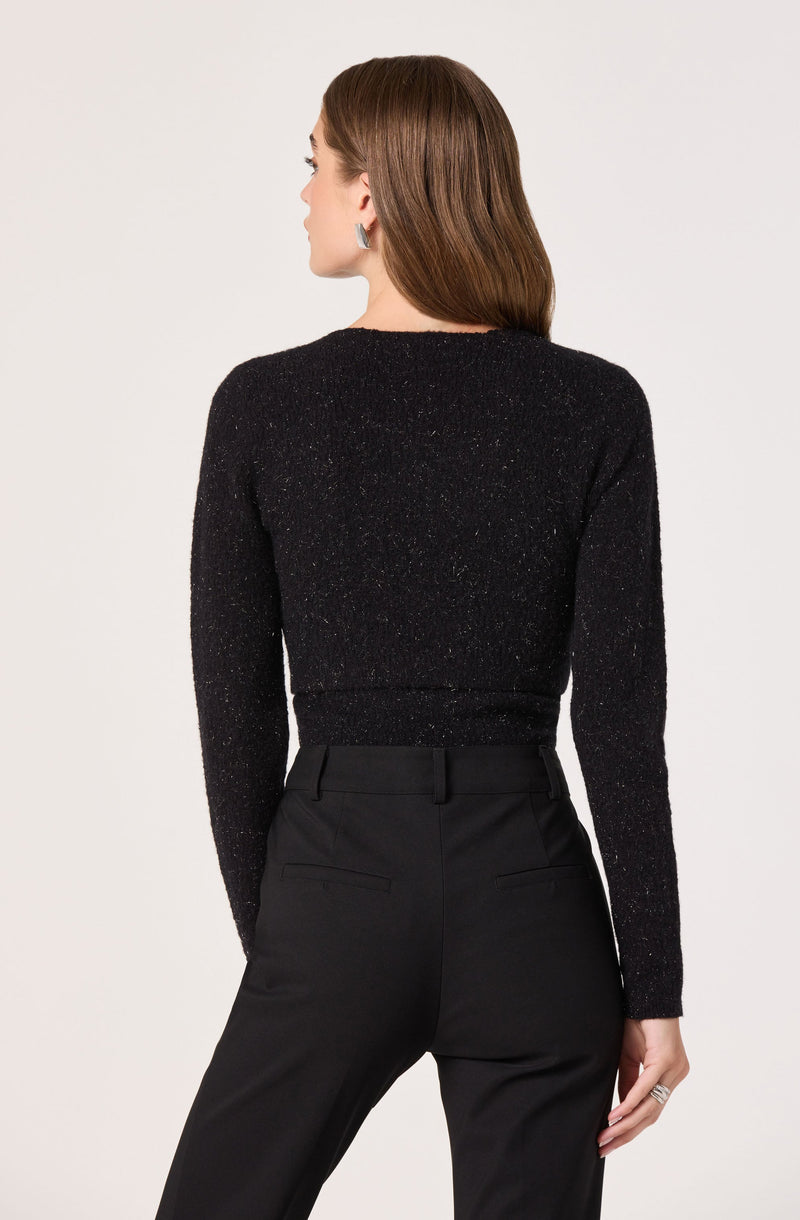A woman is wearing a black, long-sleeved, sparkly sweater with a round neckline, paired with high-waisted black tailored pants that have belt loops and back welt pockets.