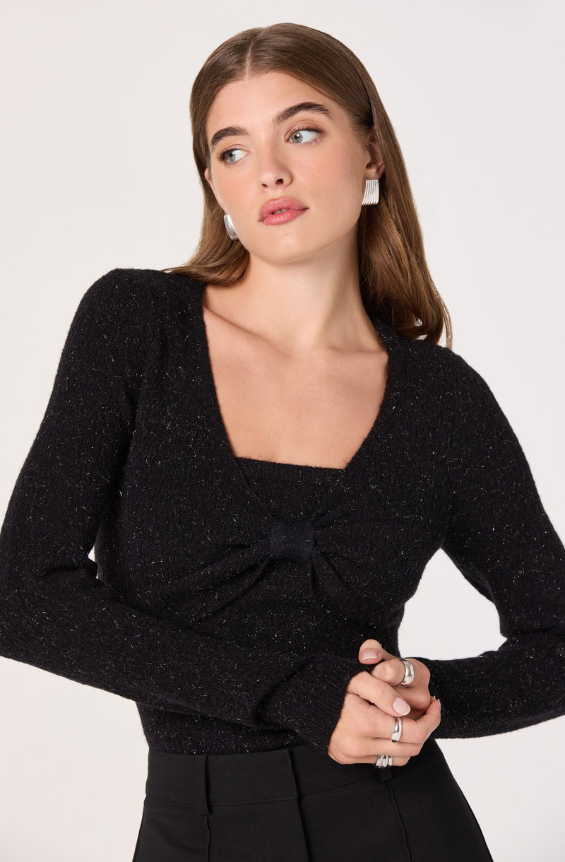 A woman is wearing a black long-sleeve top with a square neckline and a gathered detail at the bust. She is also wearing black high-waisted pants. Her accessories include large white rectangular earrings and multiple rings on her fingers.