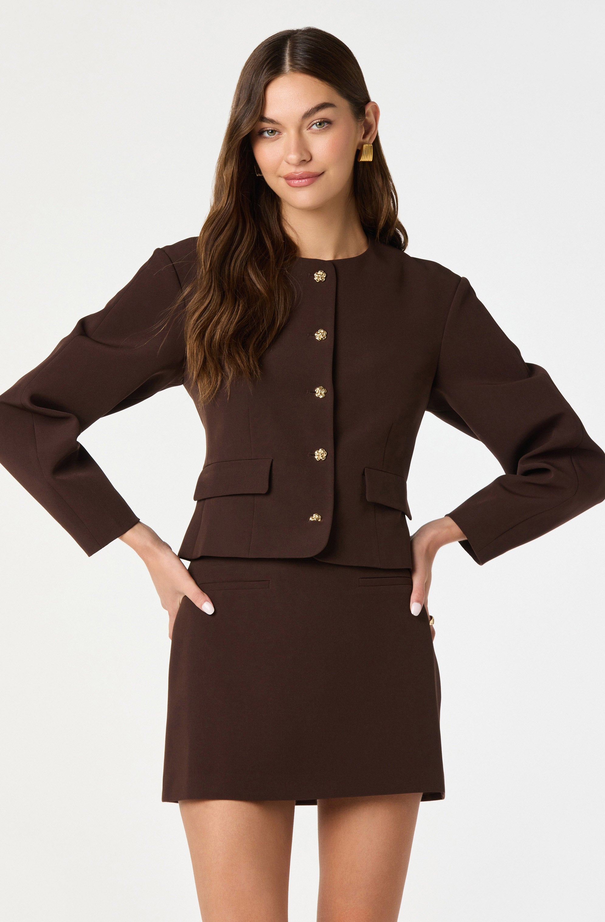 A woman is wearing a dark brown, tailored, long-sleeved blazer with a round neckline, decorated with gold buttons down the front and on the pockets. She pairs it with a matching dark brown mini skirt. She has long, wavy brown hair and is accessorized with gold earrings.