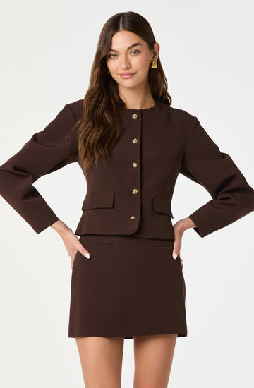A woman is wearing a dark brown, tailored, long-sleeved blazer with a round neckline, decorated with gold buttons down the front and on the pockets. She pairs it with a matching dark brown mini skirt. She has long, wavy brown hair and is accessorized with gold earrings.