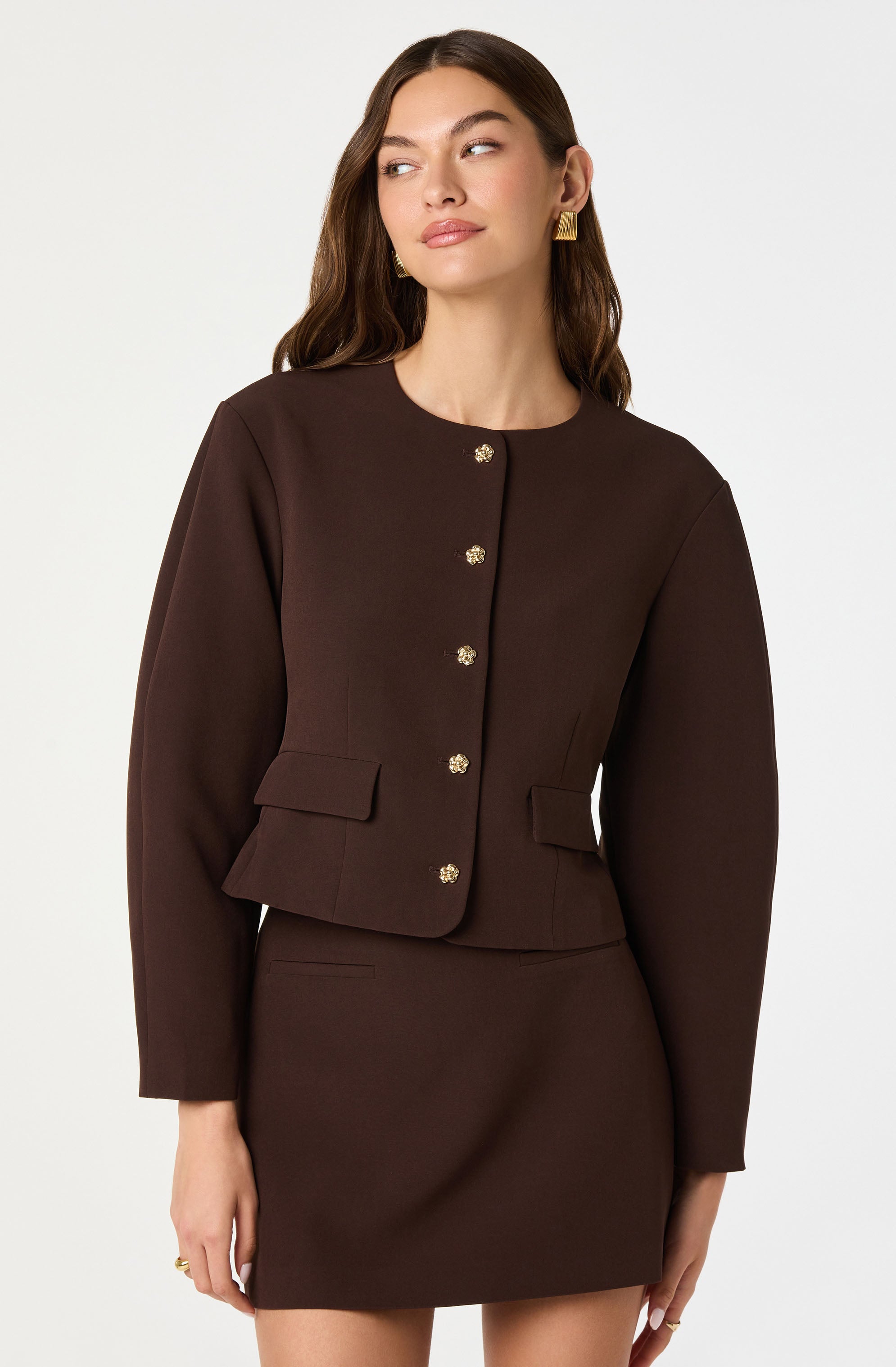 A woman is wearing a dark brown, fitted blazer with long sleeves and a matching skirt. The blazer features a rounded neckline and is adorned with gold decorative buttons down the front. The skirt is a simple, straight style that reaches mid-thigh.