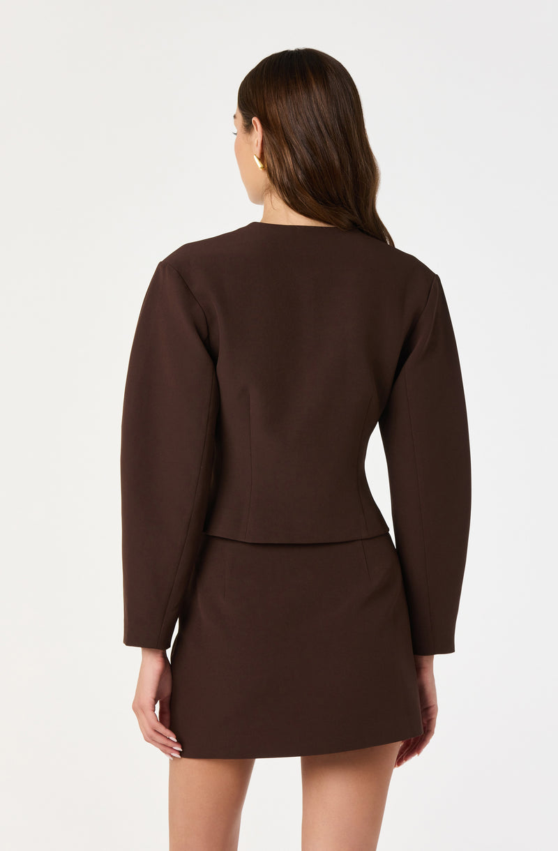 A woman is wearing a dark brown long-sleeve fitted blazer with a tailored waist, paired with a matching dark brown skirt that is above knee length.