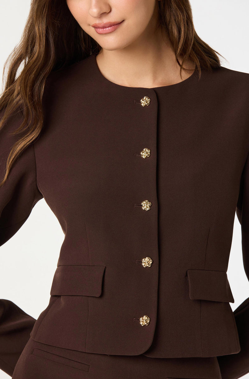A woman is wearing a tailored, dark brown blazer with a round neckline. The blazer features a row of shiny, gold-tone buttons down the front and has two decorative flap pockets at the waist.