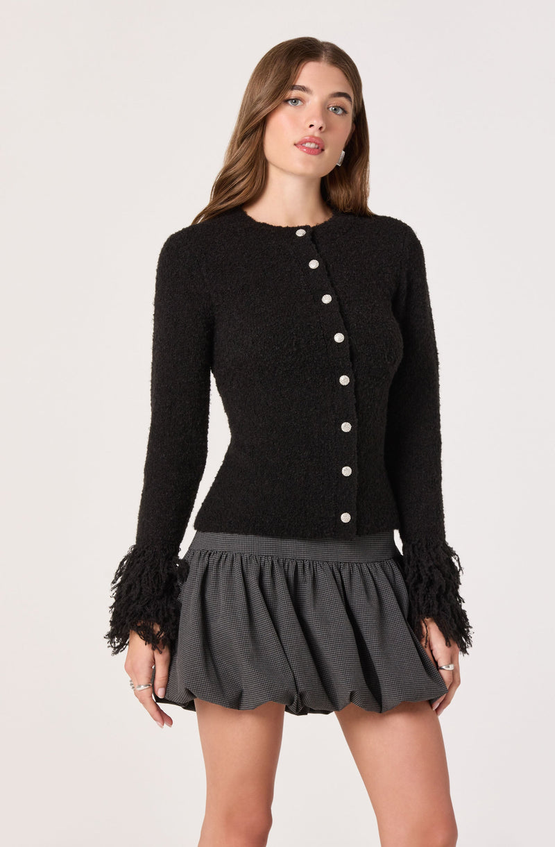 A woman is wearing a long-sleeved black textured cardigan with a round neckline and decorative buttons down the front. The cardigan features fringed detailing at the cuffs. She pairs it with a short, voluminous skirt that has a gathered, bubble-style silhouette in a dark, checkered pattern.
