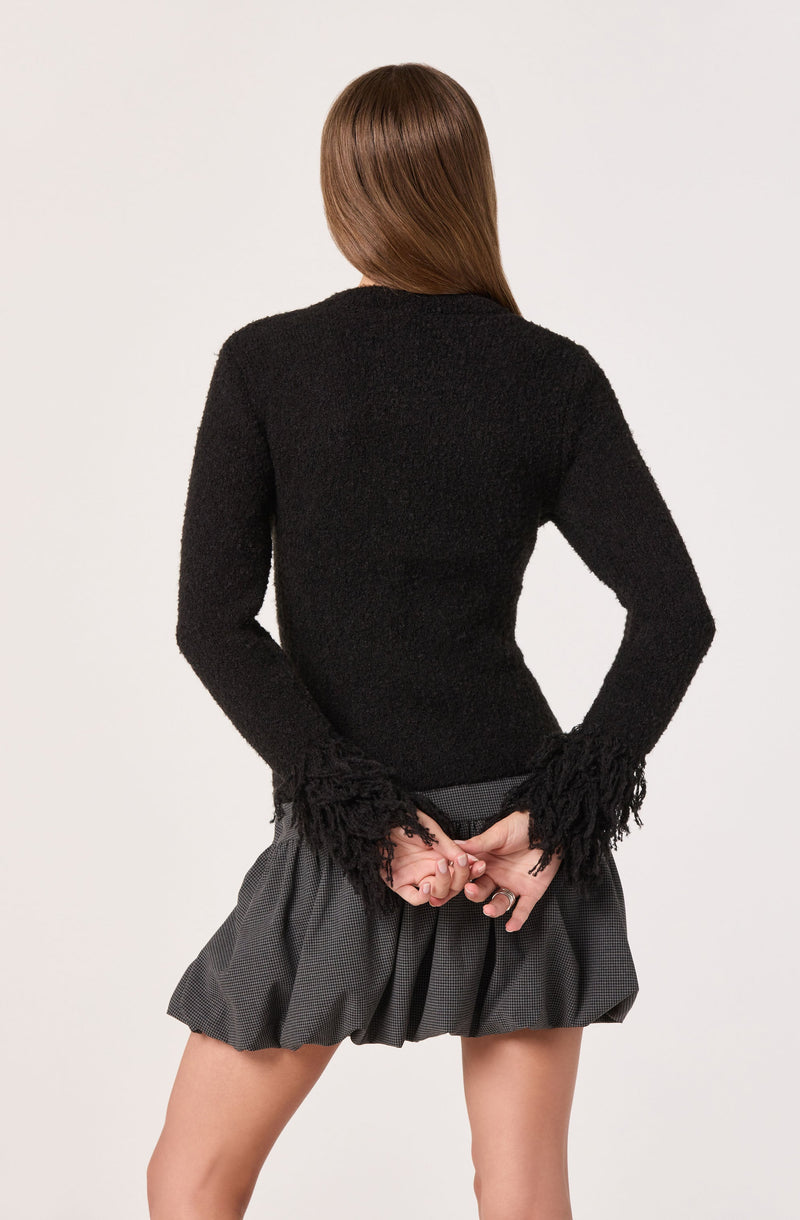 A person is wearing a black, textured sweater with long sleeves that have fringed details at the cuffs. They are also wearing a high-waisted, pleated gray skirt with a subtle checkered pattern. The person's hair is long and straight.