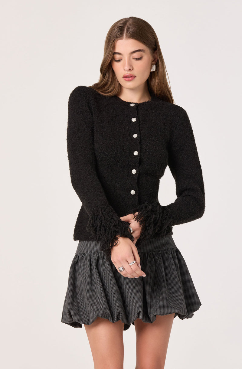 A woman is wearing a black, textured, long-sleeved cardigan with pearl buttons down the front and frayed detailing at the cuffs. She is also wearing a high-waisted, black and gray checkered, puffed skirt with a ruffled hem.