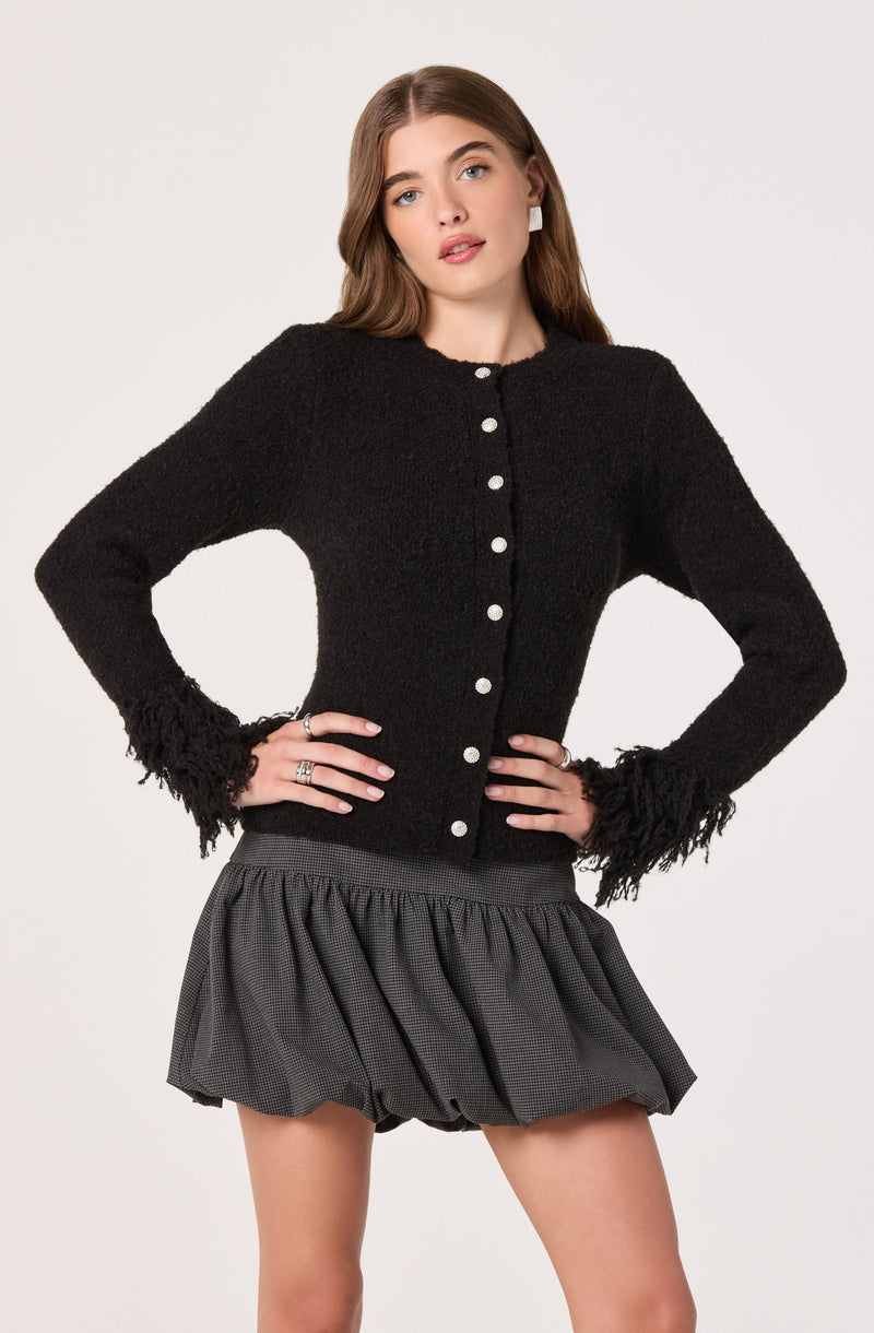 A young woman is wearing a black, textured cardigan with a round neckline and white buttons down the front. The cardigan has frayed detailing at the cuffs and hem. She pairs it with a high-waisted, gray, gathered skirt with a subtle checkered pattern.