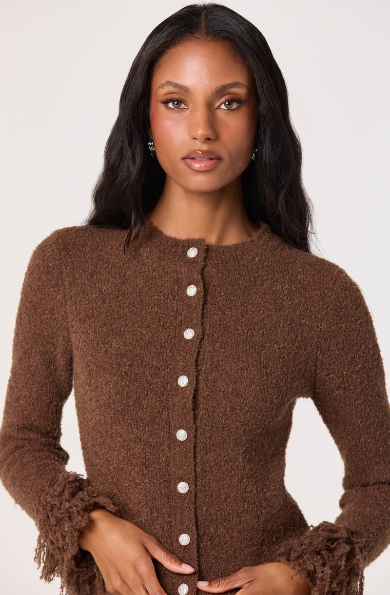 A woman is wearing a brown, textured cardigan with a fuzzy, slightly ruffled trim at the cuffs and hem. The cardigan features a row of white buttons down the front.
