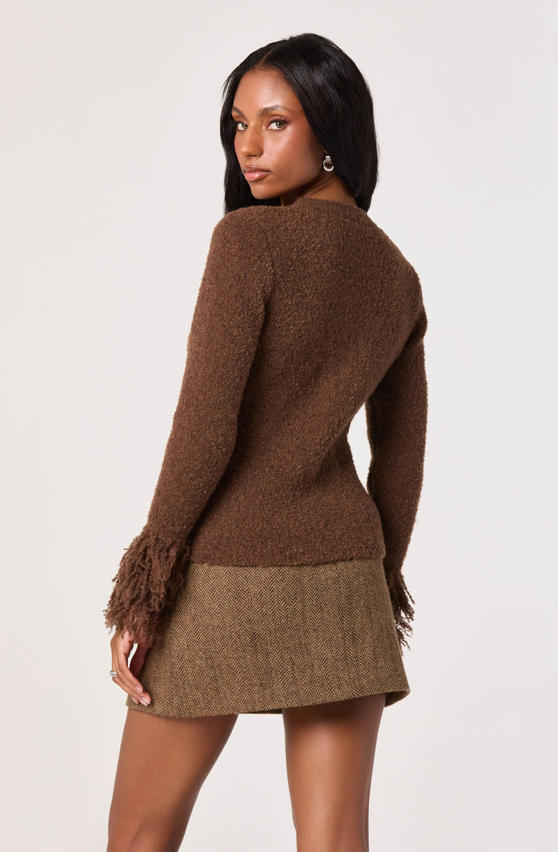 A woman is wearing a long-sleeved, brown, fuzzy sweater with textured, fringed cuffs at the wrists. She is also wearing a high-waisted, short skirt with a subtle herringbone pattern in muted earth tones.