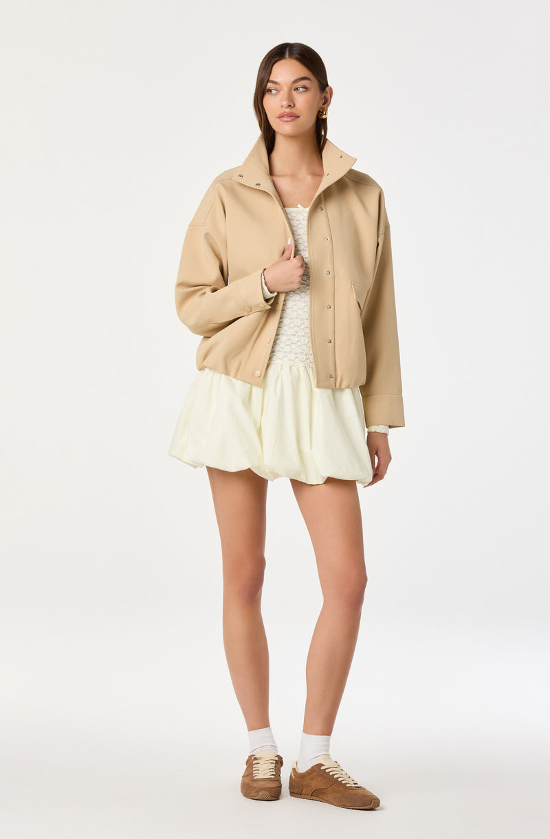 A woman is wearing a beige jacket with a high collar, long sleeves, and snap buttons down the front. Underneath, she has on a light-colored, textured top. She is also wearing a cream-colored, loose, and puffy shorts style skirt, paired with white socks and tan sneakers.