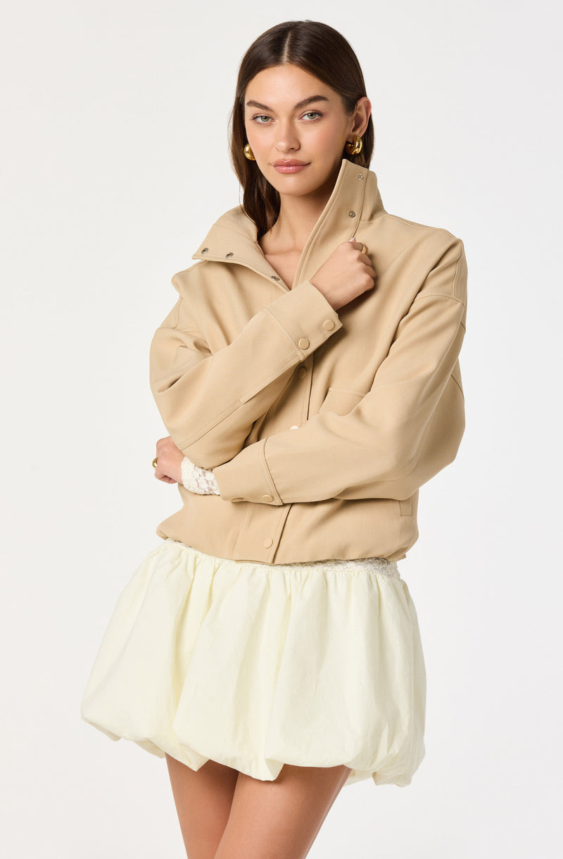 A woman is wearing a beige zip-up jacket with a high collar and long sleeves, paired with a light-colored, puffed, and gathered skirt. She has gold hoop earrings and is posing with her left arm crossed over her body and her right hand holding the collar of her jacket.