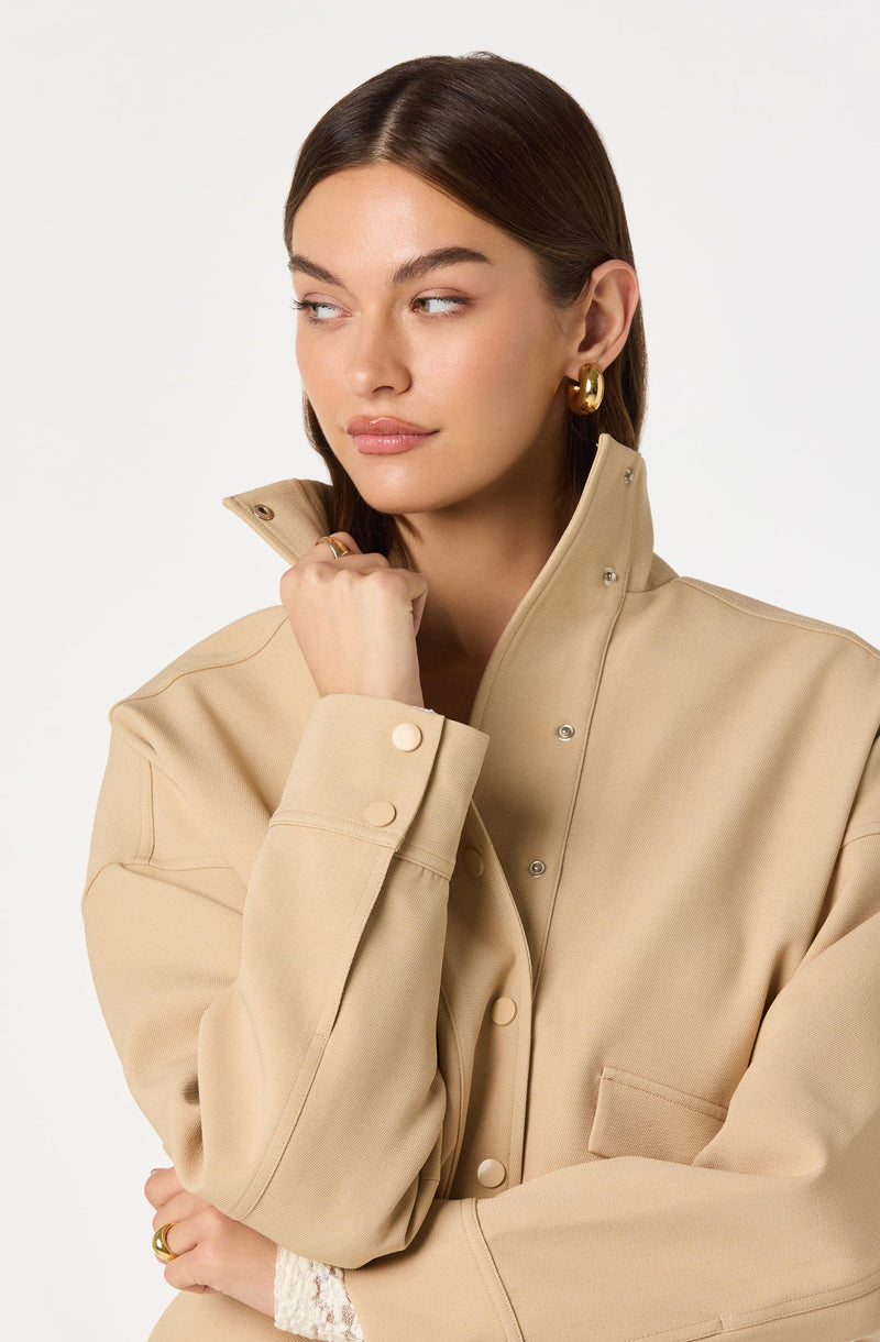 A woman is wearing a beige jacket with a high collar that is partially unzipped, featuring large buttons on the cuffs and snap closures down the front. The jacket has a structured, oversized fit with visible stitching and a pocket on the left side. She has on large, gold hoop earrings.