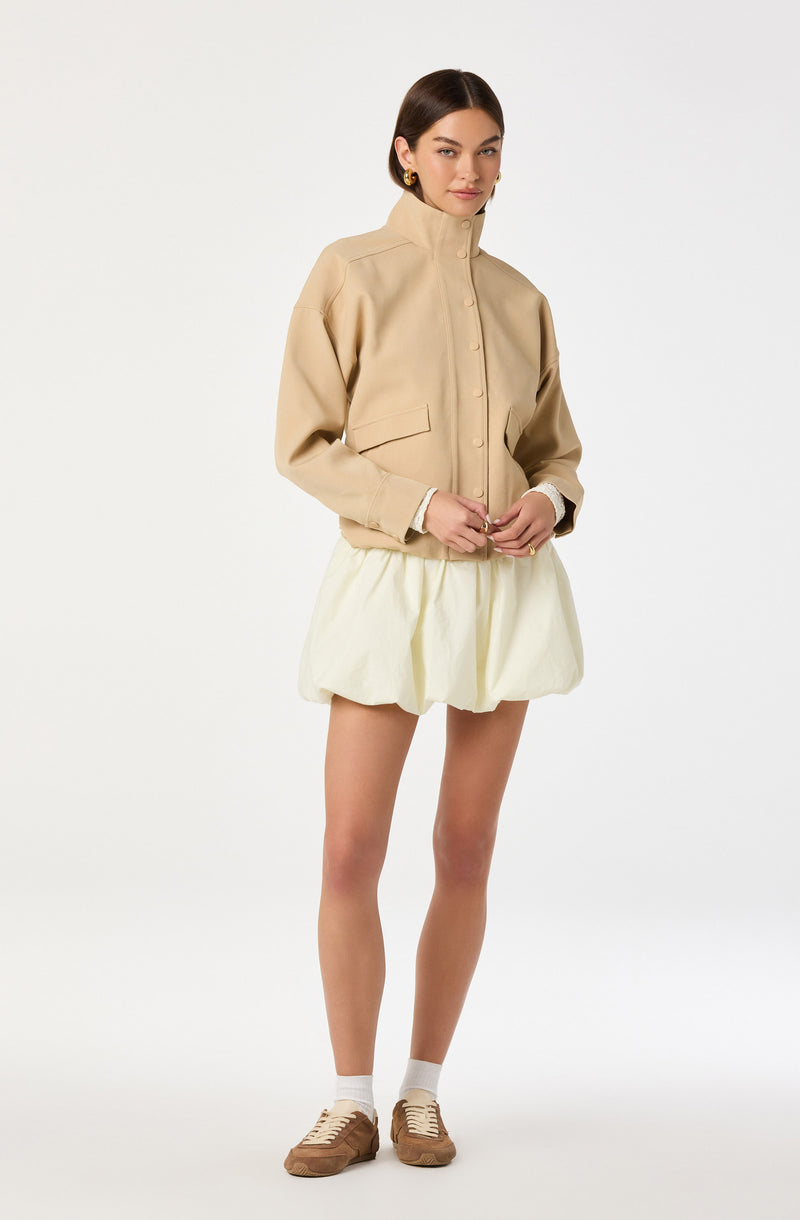 A woman is wearing a beige high-collared jacket with front buttons and long sleeves, paired with a light-colored, puffy, knee-length skirt. She has on tan sneakers with white socks and gold hoop earrings.