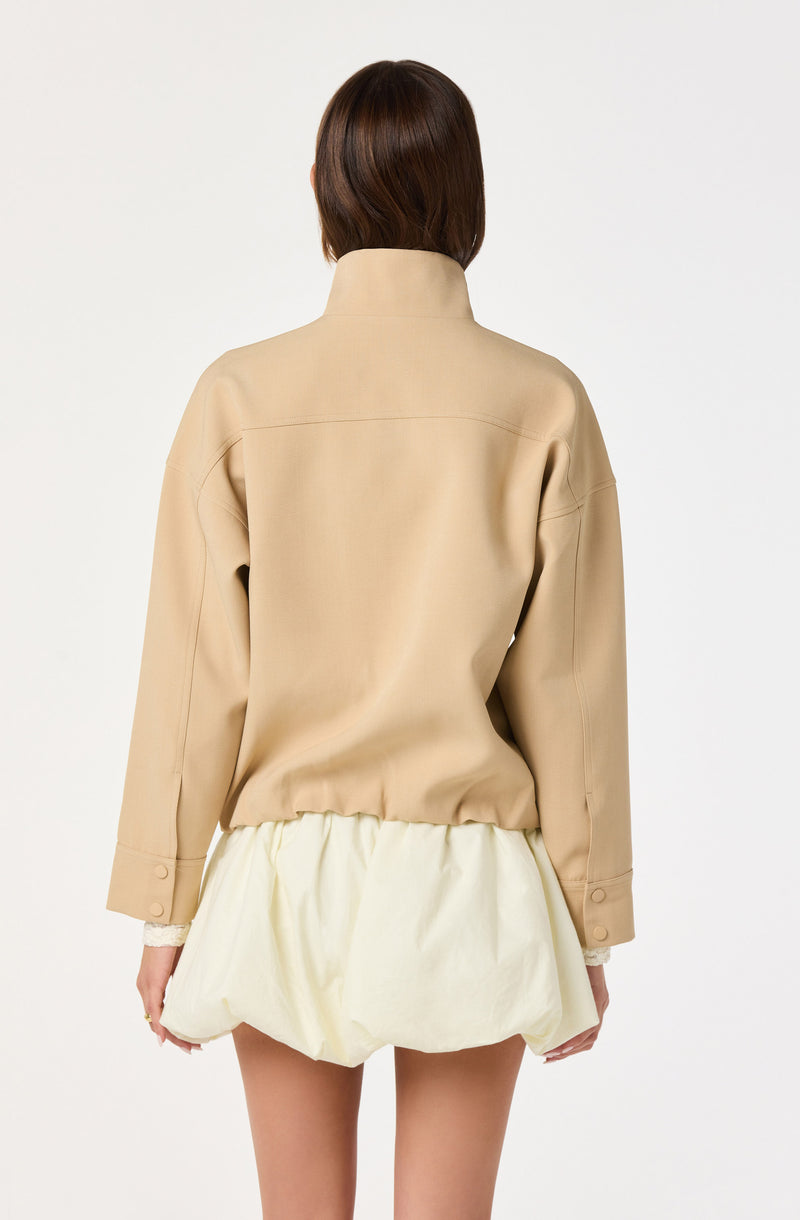 The image shows a person wearing a beige high-collar jacket with long sleeves that have buttoned cuffs, paired with a light-colored, puffy, bubble-style skirt. The person is facing away from the camera, and the background is plain white.