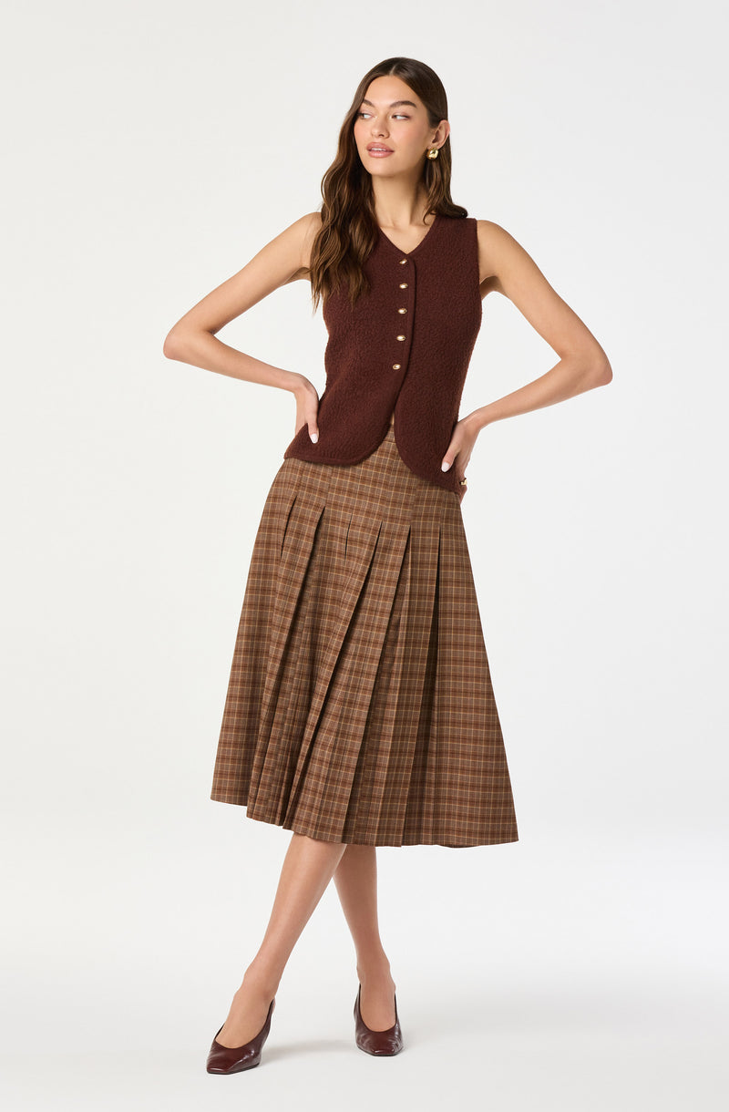 A woman is wearing a sleeveless, button-up brown vest with a textured fabric. She pairs it with a high-waisted, A-line midi skirt featuring a brown and beige plaid pattern with pleats. She completes the outfit with dark brown pointed-toe heels.