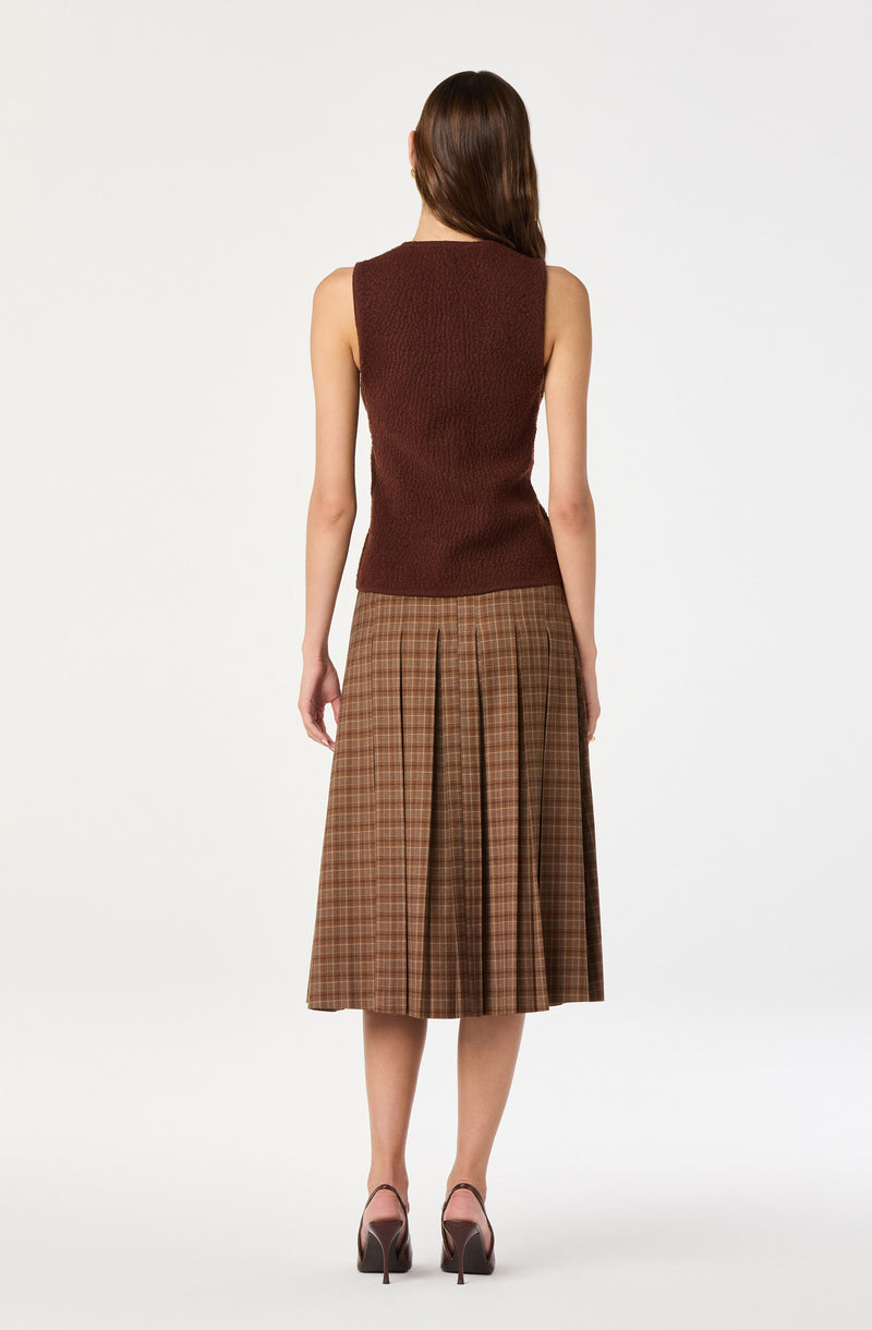 A woman is wearing a sleeveless, textured, dark red knit top paired with a high-waisted, knee-length skirt featuring a brown plaid pattern with pleats. She is also wearing dark brown high-heeled shoes.