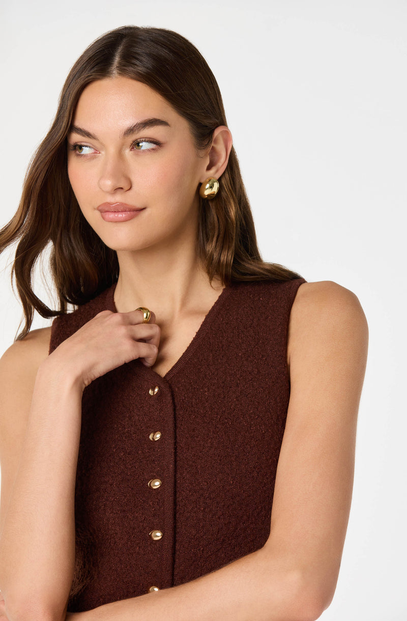 A young woman is wearing a sleeveless, dark brown textured vest with a V neckline. The vest features small, round, gold buttons running down the front. She accessorized with large, shiny gold stud earrings and a gold ring on her right hand.