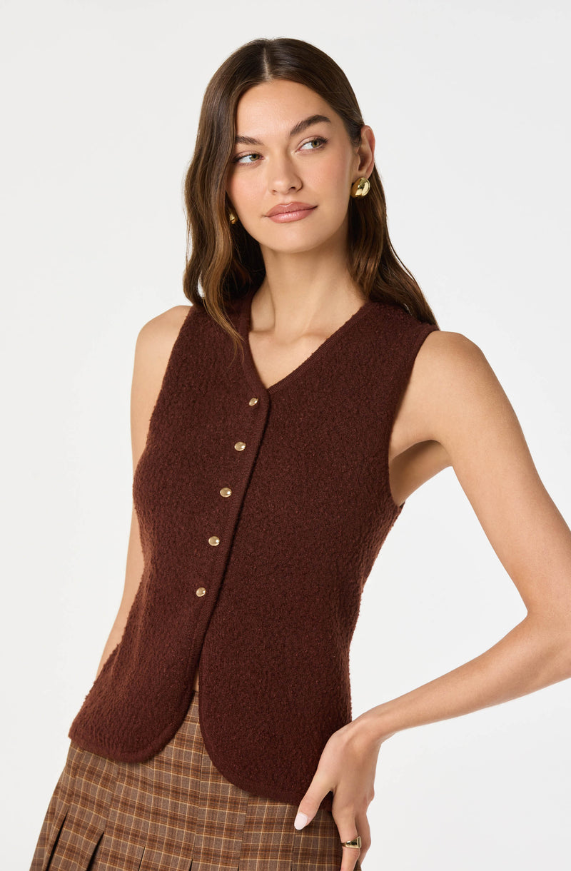 A woman is wearing a sleeveless, textured, deep brown vest with a V-neckline and gold buttons running down the front. She is also wearing a pair of gold hoop earrings and appears to have a plaid skirt with brown and beige tones.