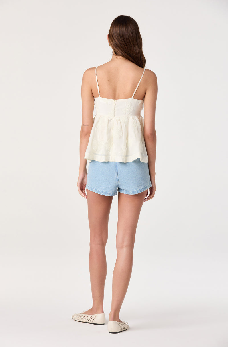 Woman wearing a cream-colored, sleeveless, strappy top and light blue denim shorts.